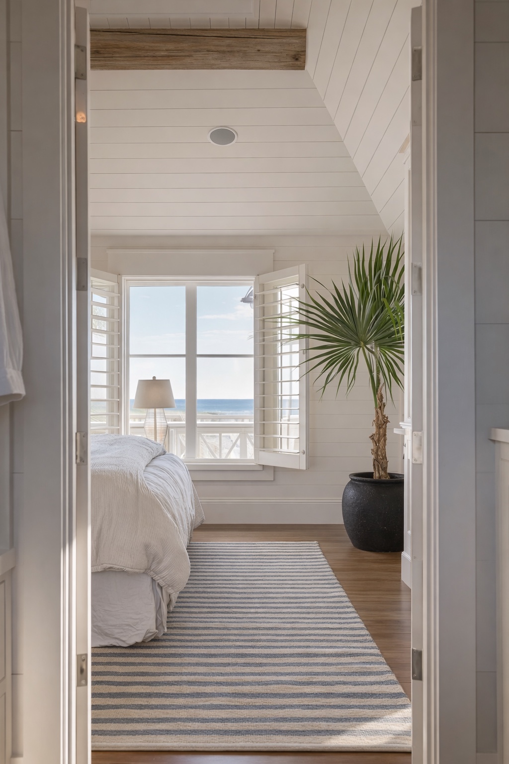 Coastal bedroom view with striped rug, shiplap ceiling, and bright ocean-facing window.