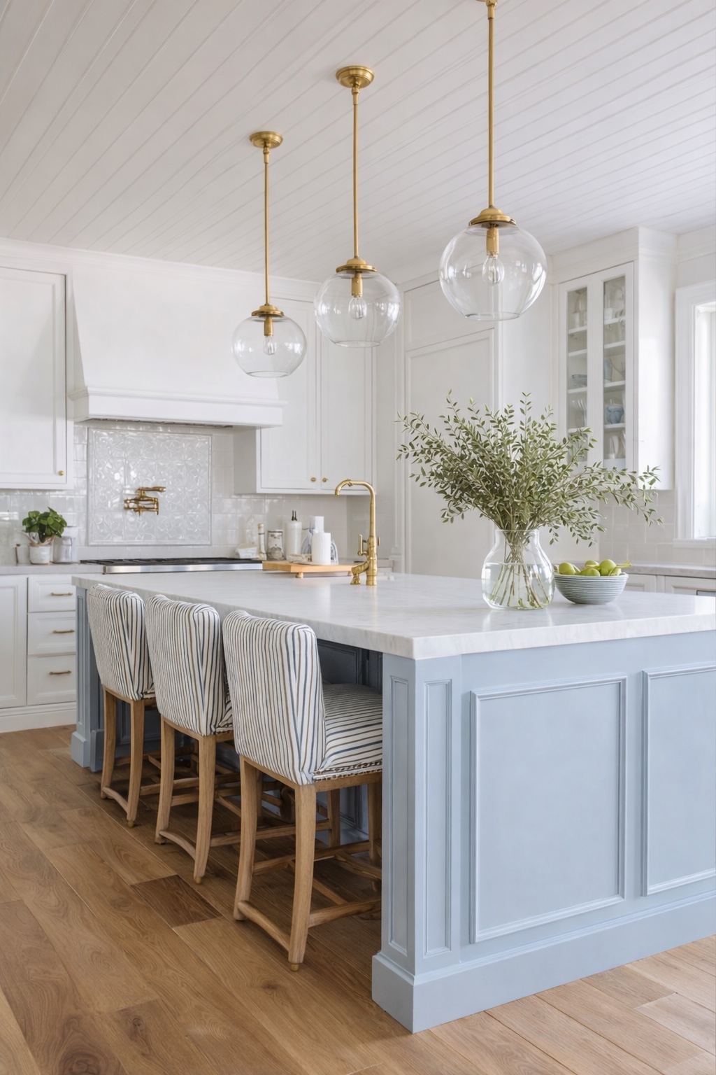 White kitchen with soft blue island, brass pendants, striped stools, and light wood floors.